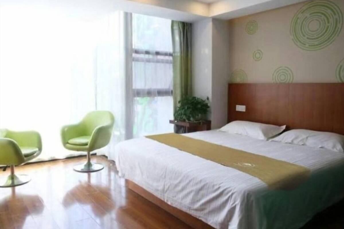 A hotel room with a bed, two green chairs, a window with curtains, and a wall with circular patterns.