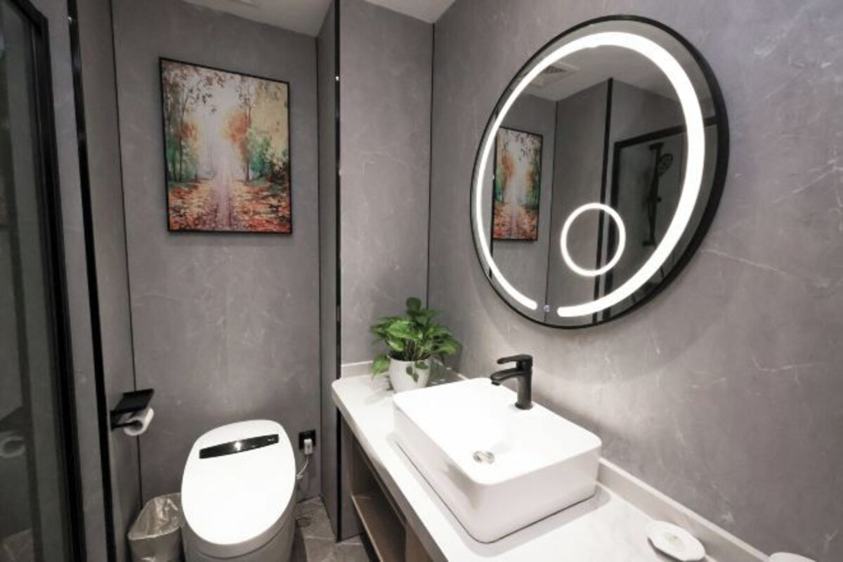 A modern bathroom with a white sink, a round mirror with a light, and a painting on the wall.