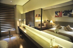 Habitación de hotel con un banco largo, dos camas, un escritorio y un espejo grande.
