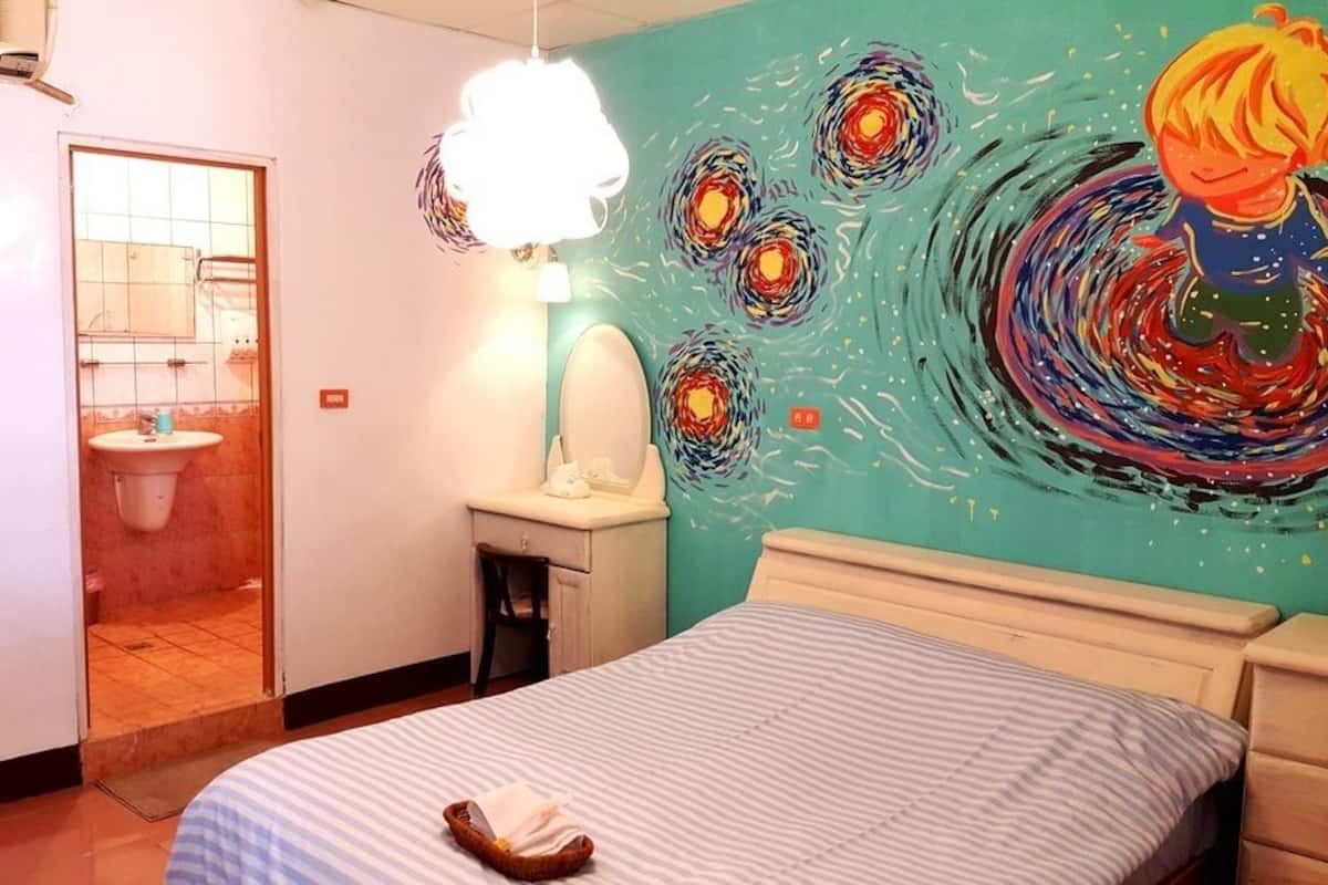 Una habitación con cama, una mesita de noche, una pared con un cuadro colorido, una puerta de baño y una lámpara colgante.