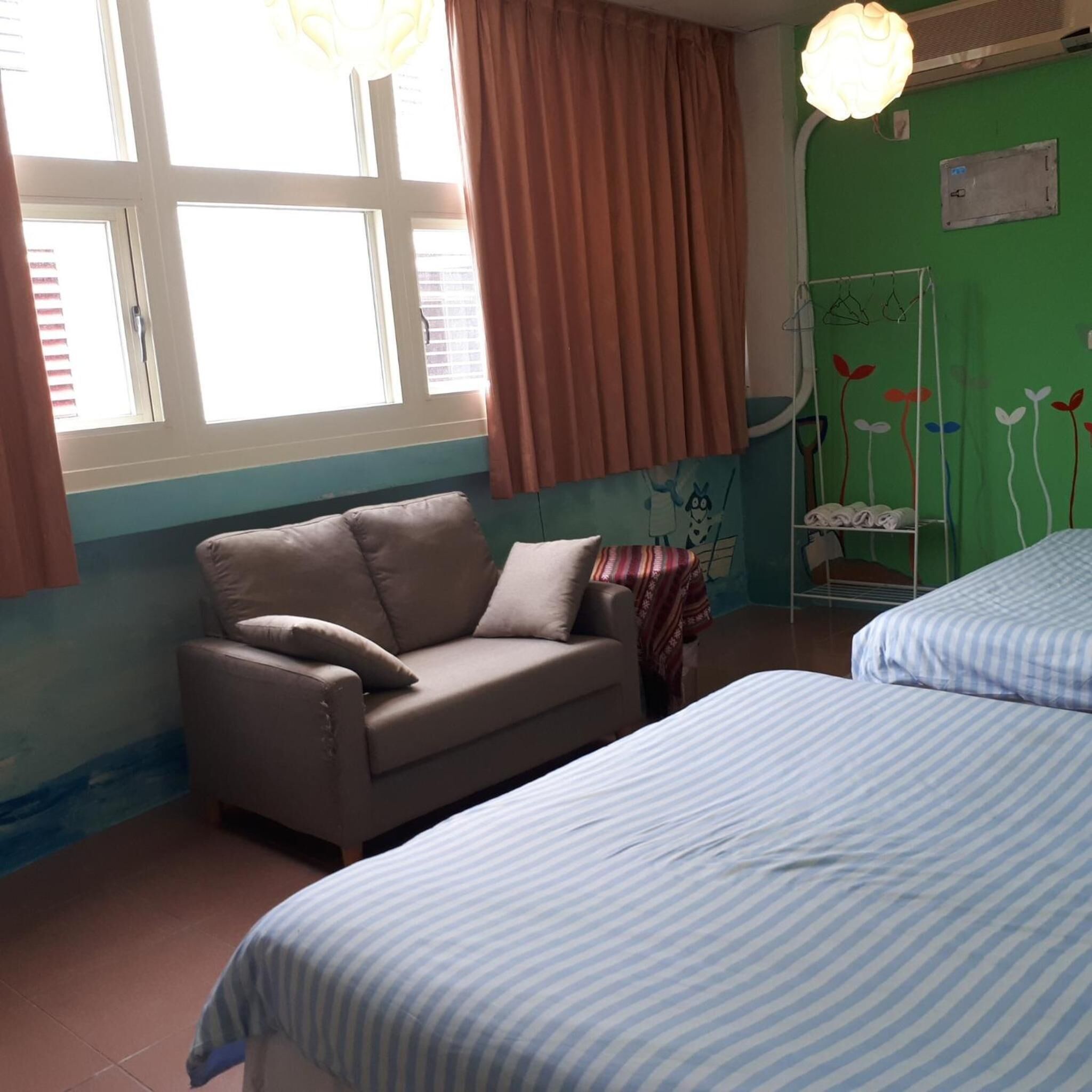 A room with two beds, a sofa, a window with curtains, and a green wall with a floral mural.