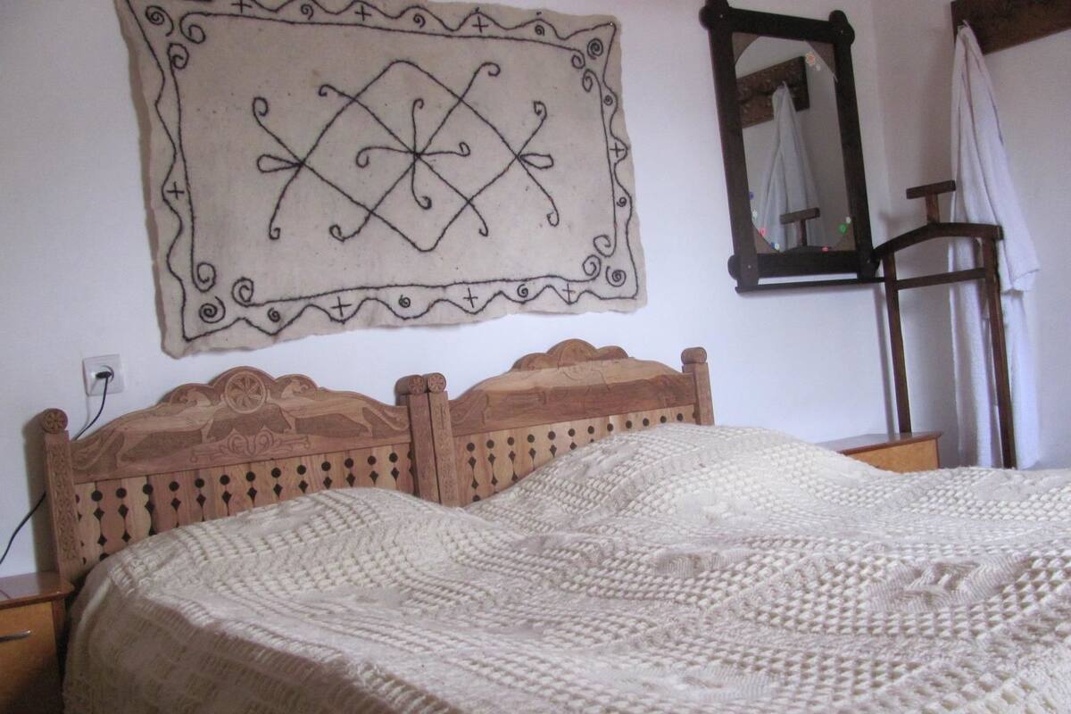 A bed with a wooden headboard, a white knitted blanket, a wall tapestry, and a mirror.