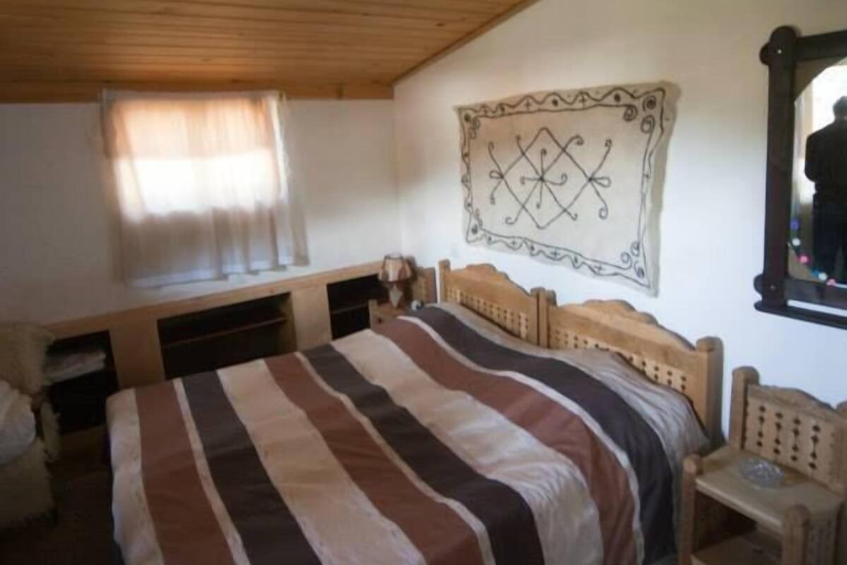 A bedroom with a wooden ceiling, a large bed with striped bedding, a bedside table, a chair, and a wall-mounted artwork.