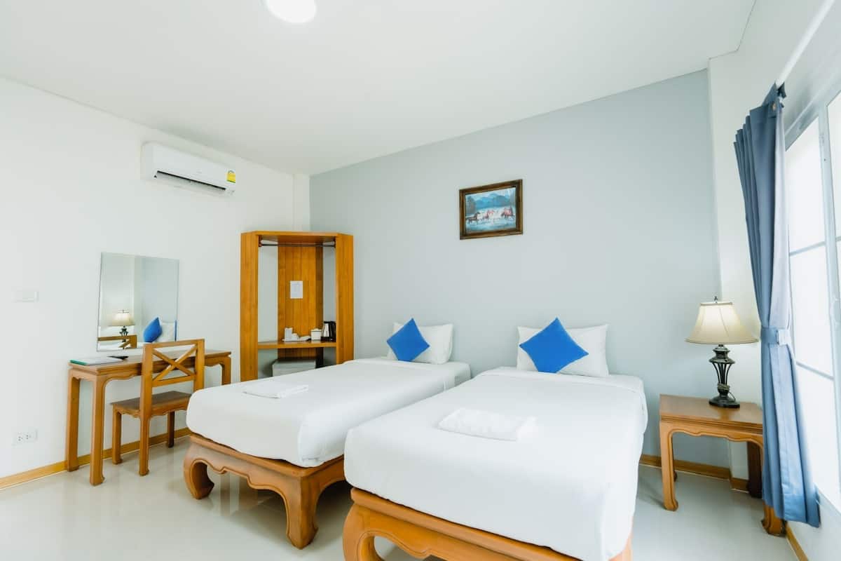 Habitación de hotel con dos camas individuales, un escritorio con silla, un espejo, un equipo de aire acondicionado, una mesita de noche con lámpara y una ventana con cortinas.