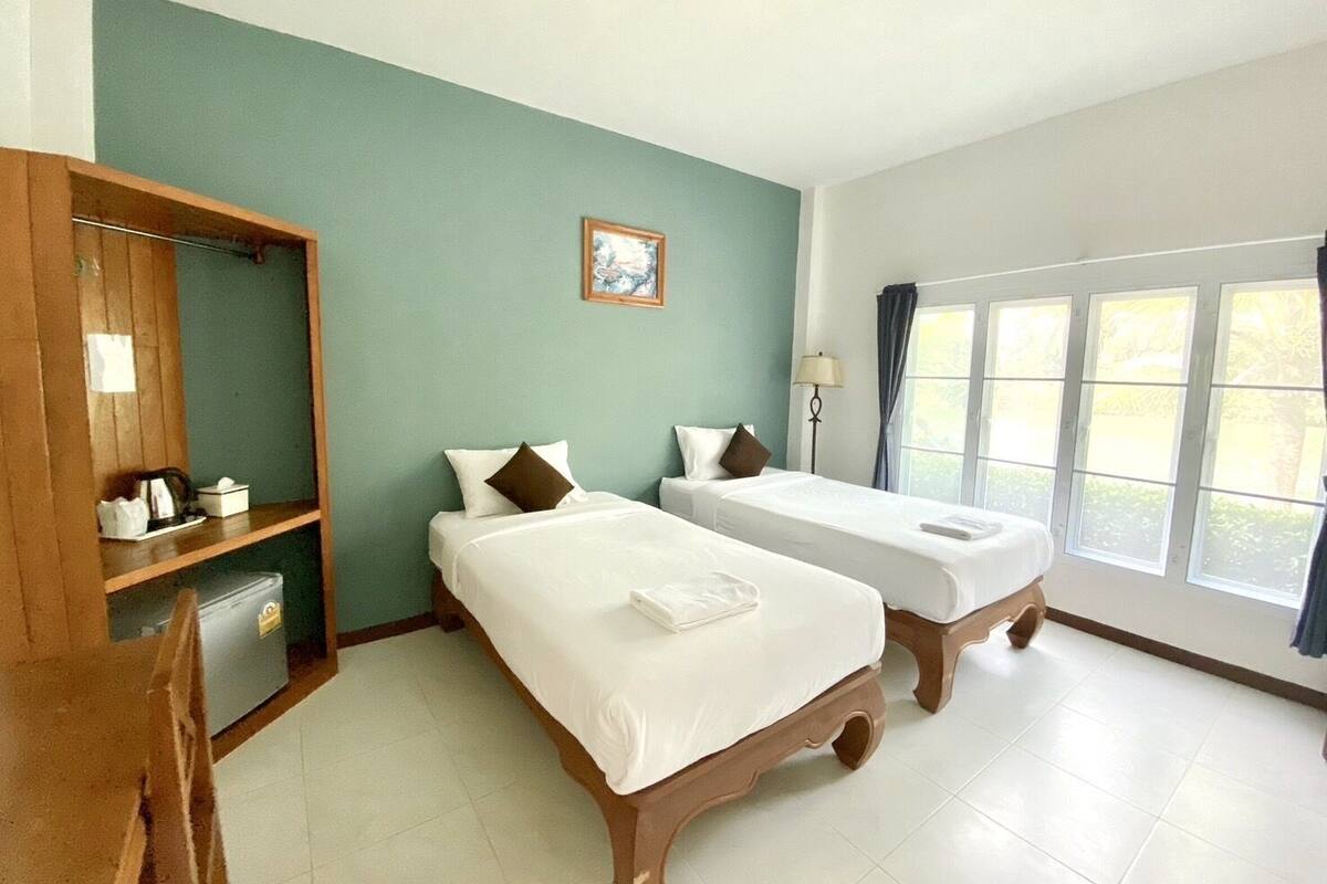 Habitación de hotel con dos camas individuales, un armario de madera, un escritorio con silla, una ventana con cortinas y un pequeño cuadro en la pared.