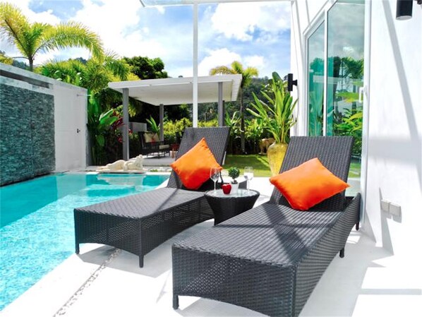 A modern outdoor pool area with two lounge chairs and a small table, surrounded by lush greenery and a clear blue pool.