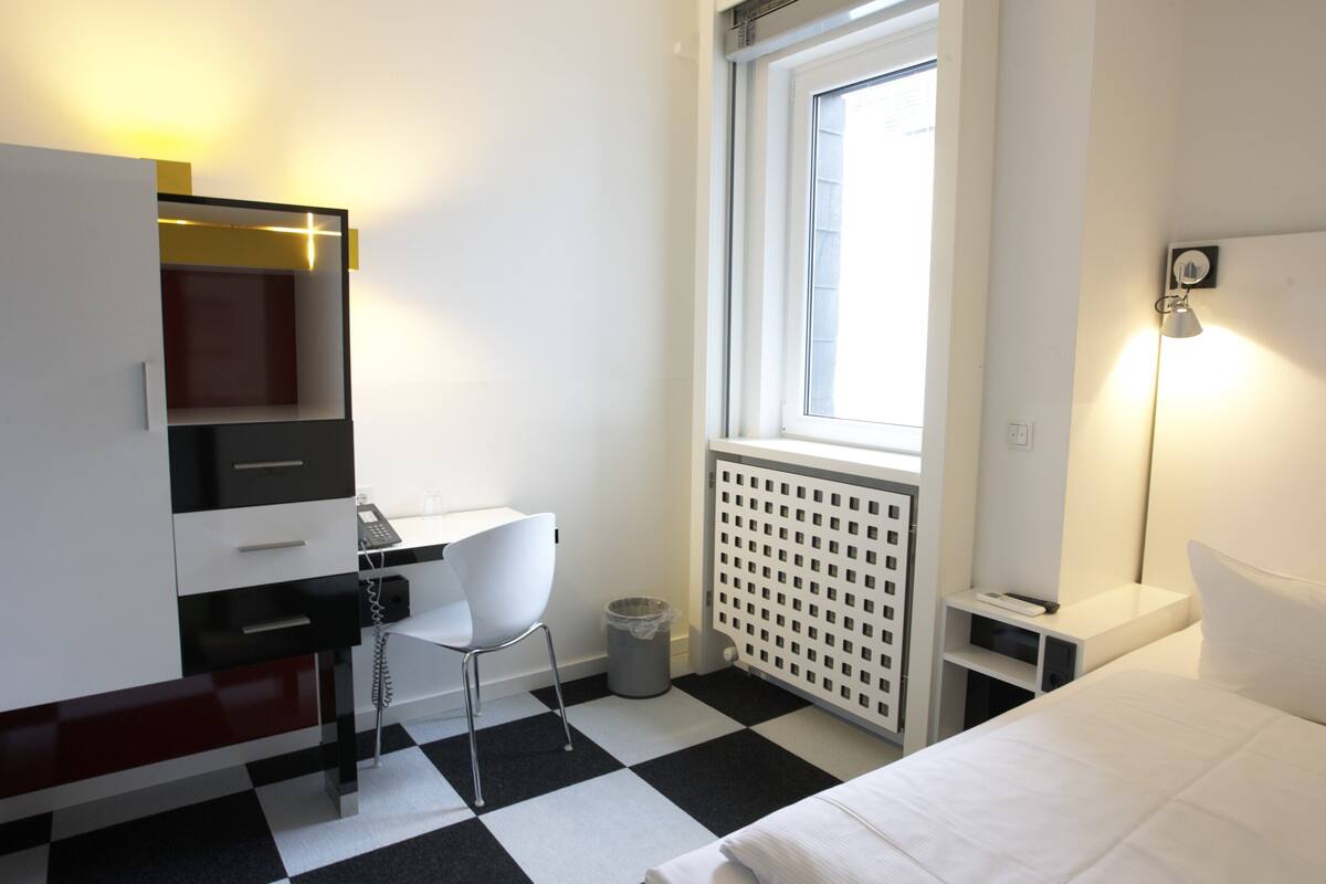 Een hotelkamer met een zwart-wit geruit vloerkleed, een bureau met stoel, een bed, een radiator en een raam met jaloezieën.