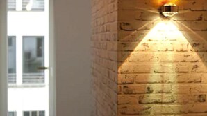Een gang met een bakstenen muur en een deur die leidt naar een lichte kamer.