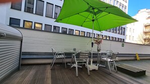 Een terras met metalen stoelen en een tafel, een groene parasol en een bloemenvaas.