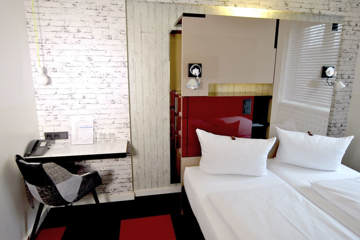 Een hotelkamer met een witte bakstenen muur, een bureau met telefoon, een stoel, een bed met witte beddengoed en een rode accentmuur.