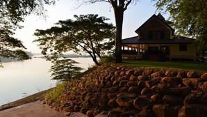A lakeside property with a stone wall, a large tree, and a yellow building with a porch. - Wopakok Hotel (Pakse)