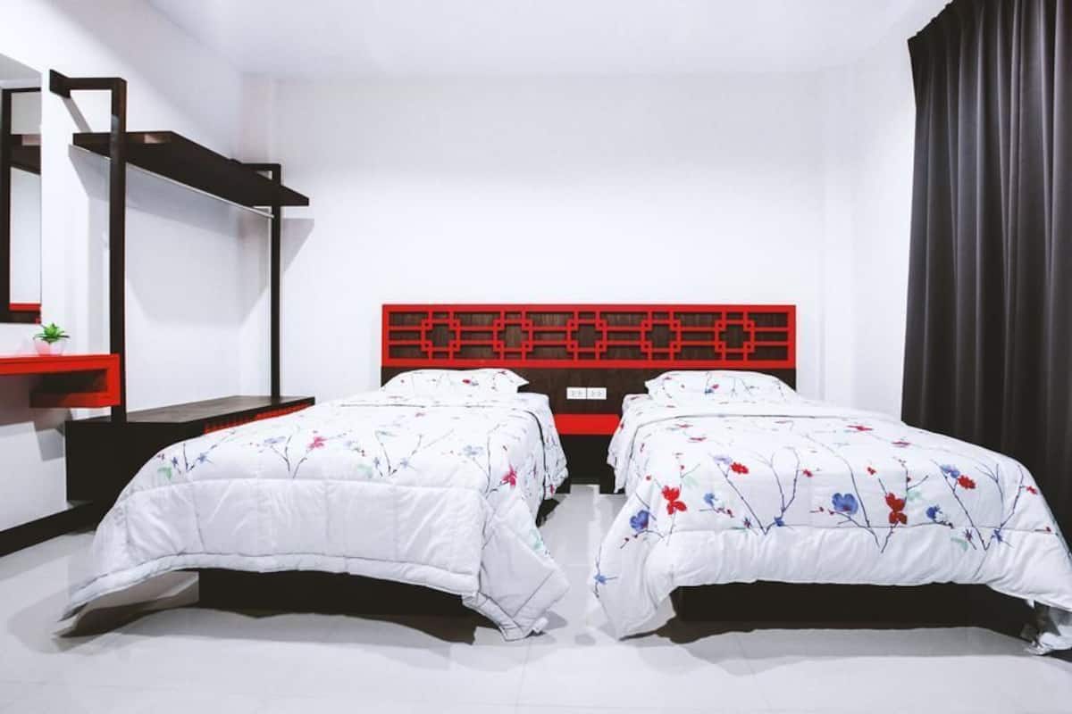 A hotel room with two beds, a red headboard, a small desk with a plant, and a black curtain.