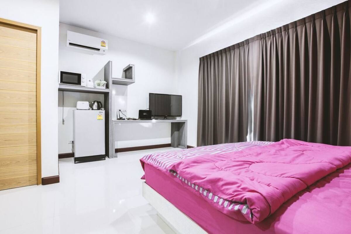 A hotel room with a bed, a desk with a computer and a television, an air conditioning unit, and a window with curtains.