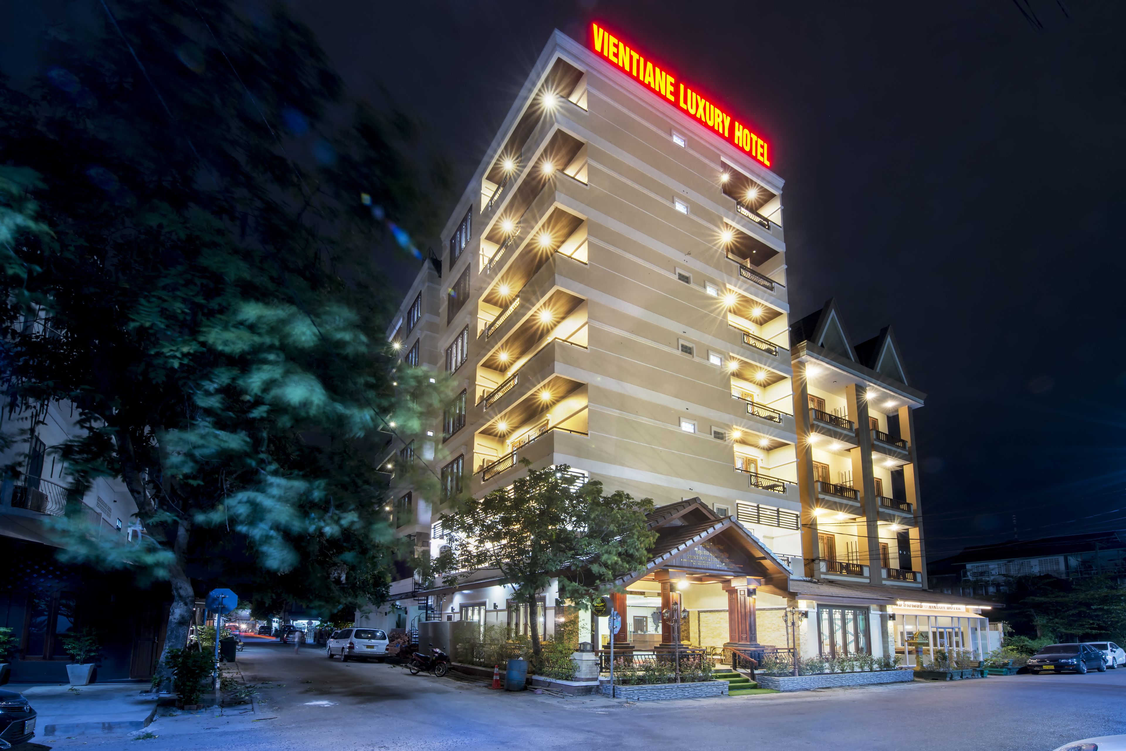 Un hotel de varios pisos con un letrero llamativo que dice "VIENTIANE LUXURY HOTEL" en la parte superior. El edificio cuenta con balcones en cada piso, una entrada cubierta y está rodeado de árboles y coches estacionados.