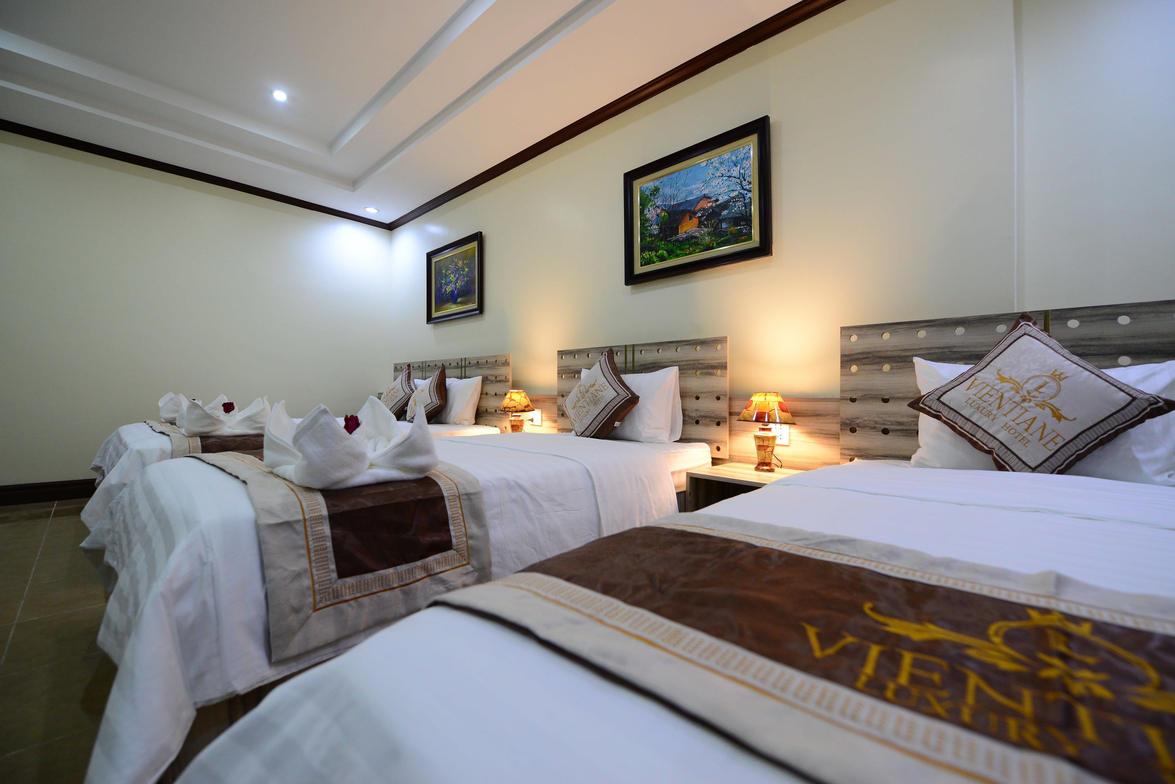 Habitación de hotel con dos camas, cada una con una almohada y una manta. Hay dos mesitas de noche con lámparas. La habitación tiene un techo con iluminación empotrada y un cuadro en la pared.