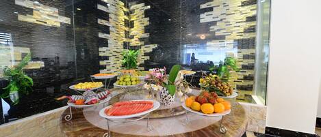 Un bufé con diversas frutas y verduras, un florero decorativo, y un diseño interior moderno con una pared de vidrio y un techo estampado.