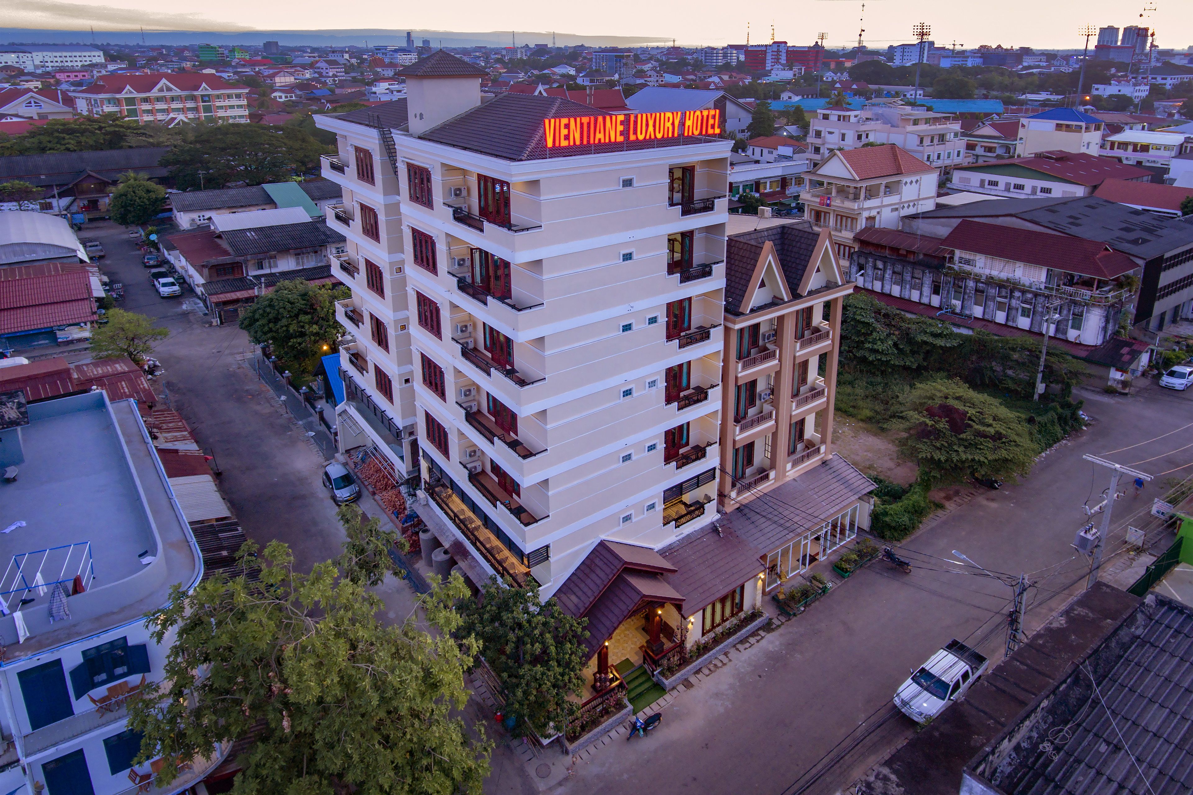 Egy többemeletes épület, amelynek erkélyei vannak, az elején egy „VIENTIANE LUXURY HOTEL” feliratú tábla, és előtte egy parkolóhely található.