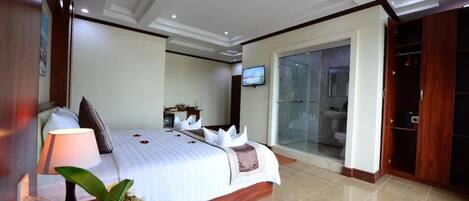 A hotel room with a large bed, a TV, a bathroom with a glass shower, and a wooden wardrobe.