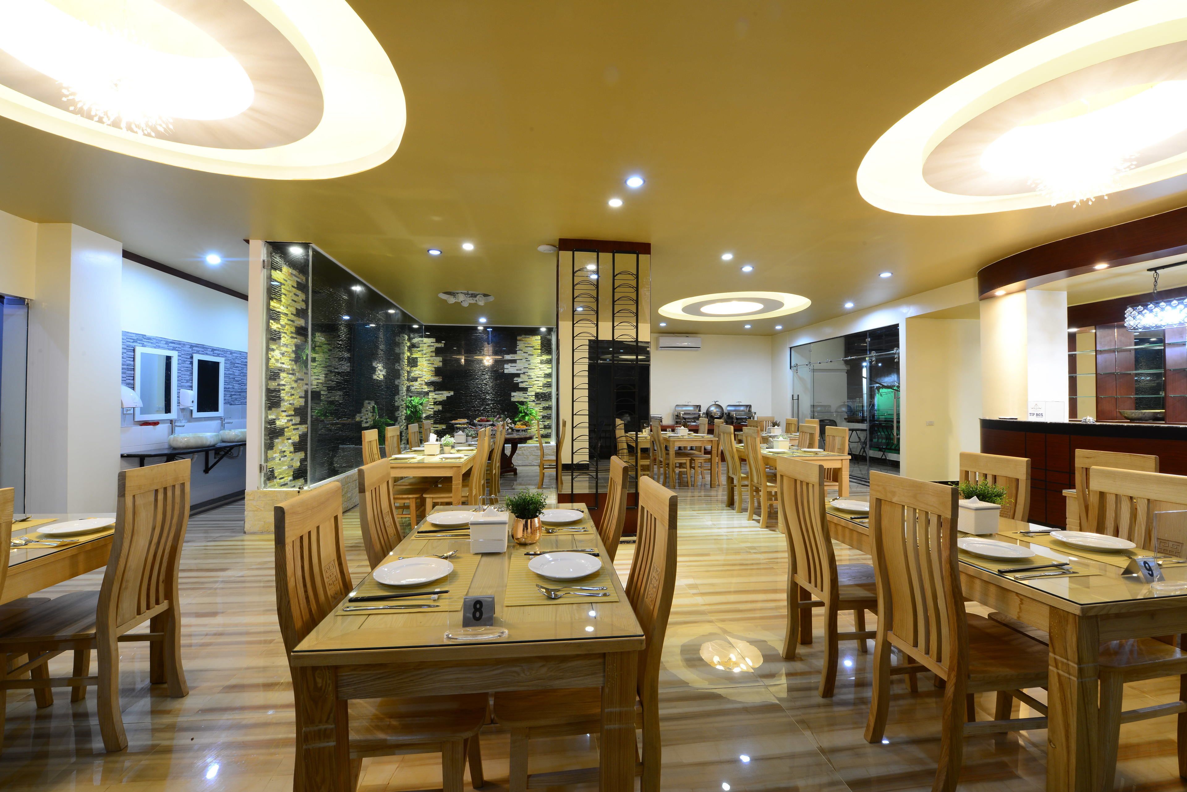 A spacious dining area with wooden tables and chairs, modern lighting fixtures, and a view of the kitchen in the background.