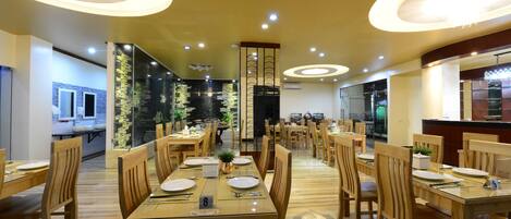 A spacious dining area with wooden tables and chairs, modern lighting fixtures, and a view of the kitchen in the background.