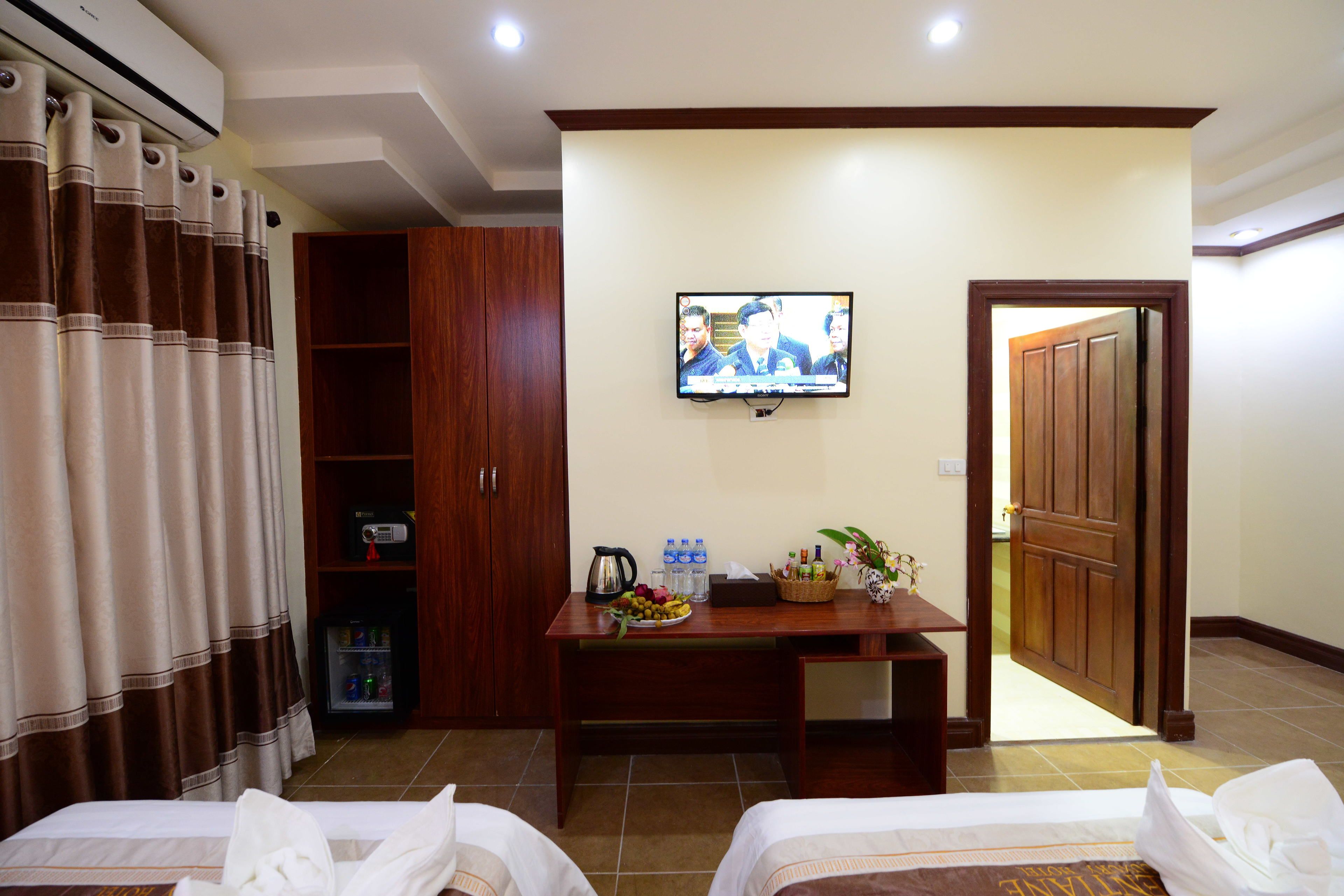 A hotel room with two beds, a TV on the wall, a wooden cabinet, a table with a fruit basket, and a door leading to another room.