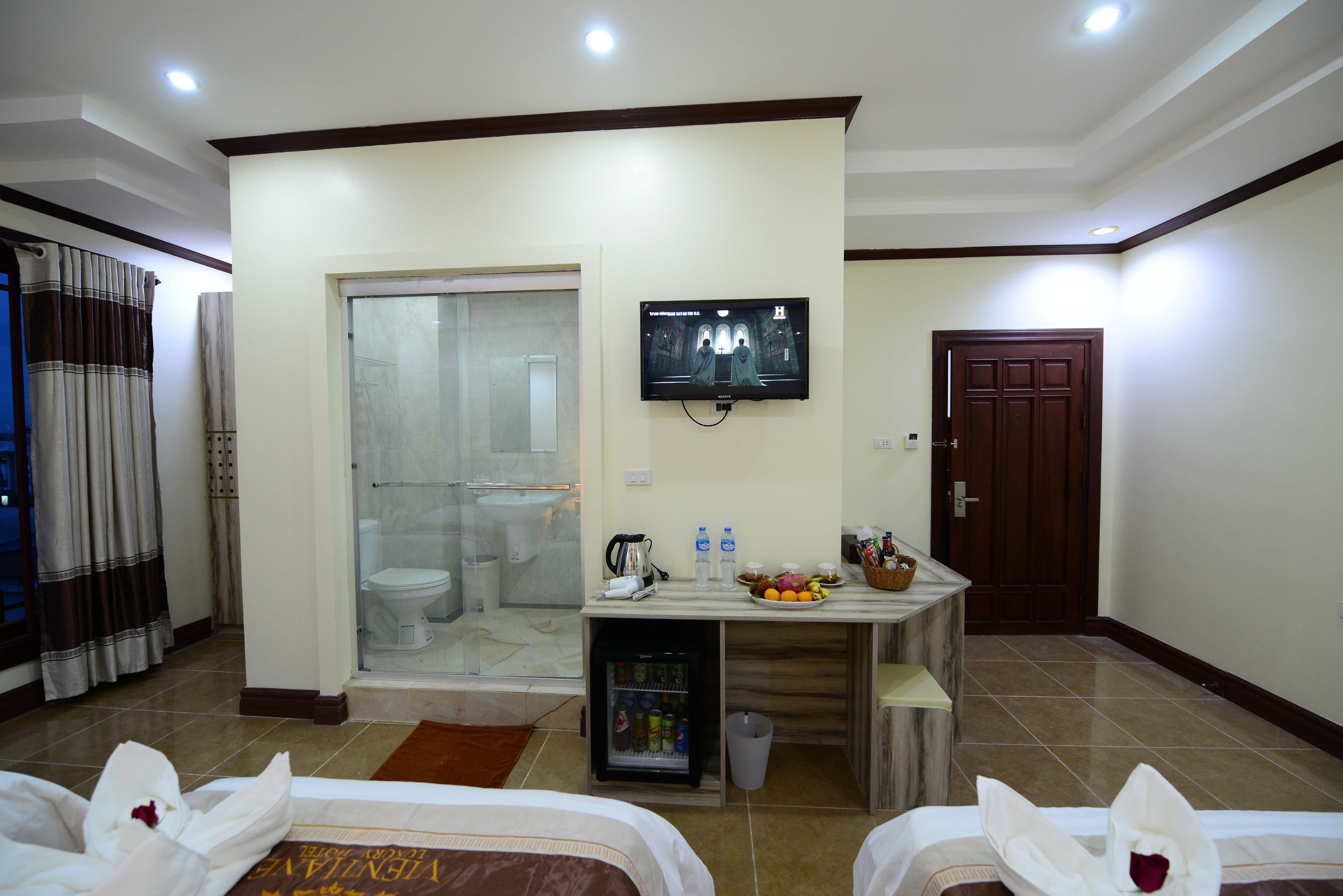 A hotel room with a bed, a small table with a TV, a mini-fridge, and a bathroom visible through a glass door.