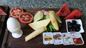 Une planche en bois sur laquelle sont disposés divers aliments, notamment des tomates en tranches, de la pastèque, du fromage, des olives et des en-cas emballés.