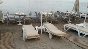 Un espace aménagé au bord de la plage, avec des chaises longues et des tables, un sol sablonneux et une vue sur la mer et les collines lointaines.