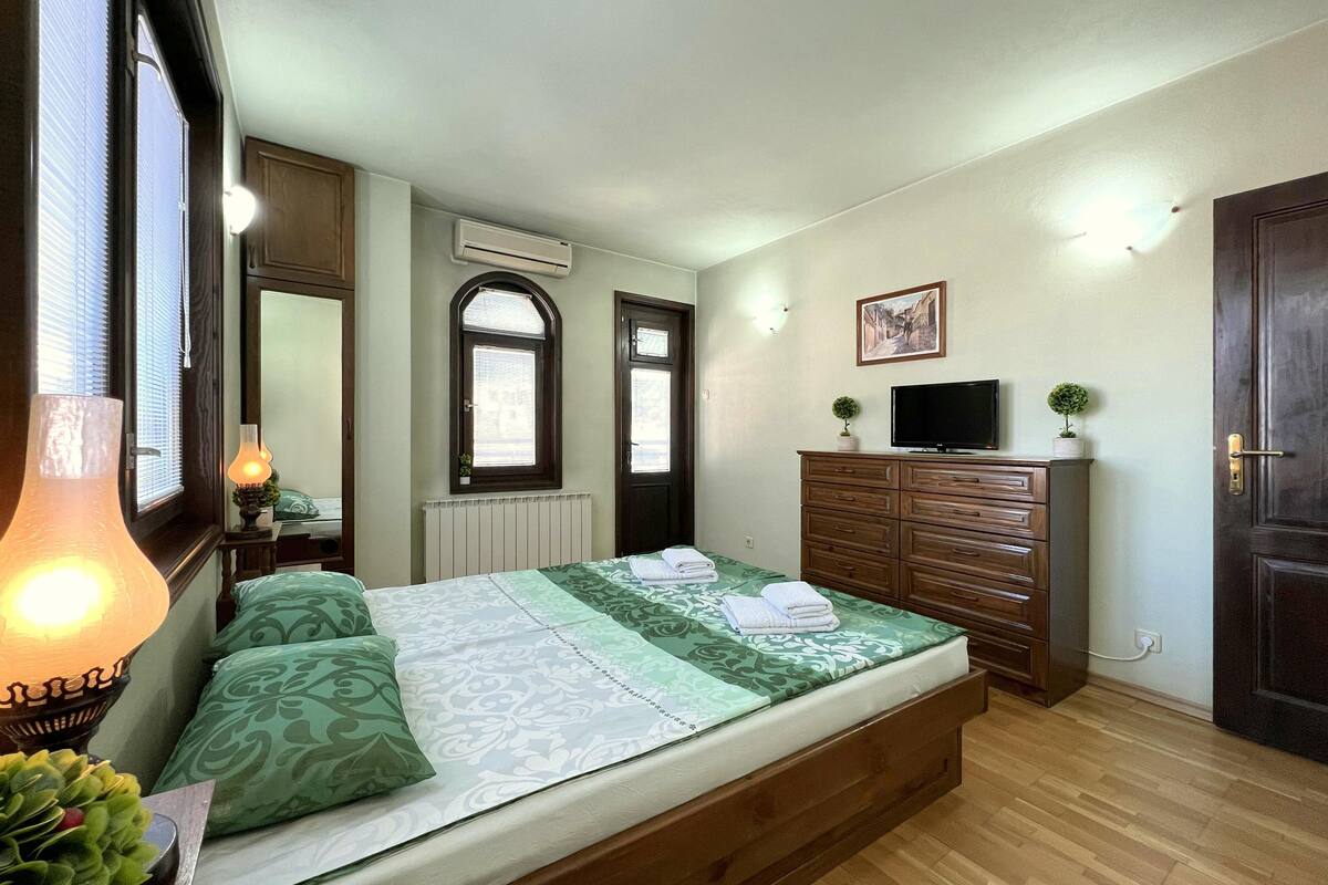 A bedroom with a bed, dresser, TV, and a window with blinds.