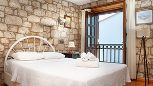 Un dormitorio con pared de piedra, puerta de madera, cama con ropa blanca, mesita de noche, lámpara y vista al mar.