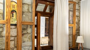Una habitación con paredes de piedra, ventanas de madera y un mesita de noche negra con una lámpara, y vista a la calle.