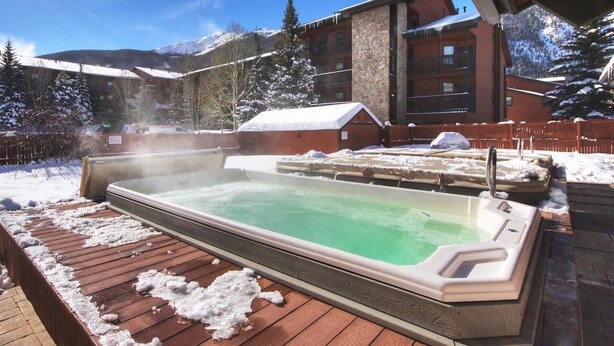 A hot tub on a wooden deck with snow around it, set against a backdrop of a mountain and a building.