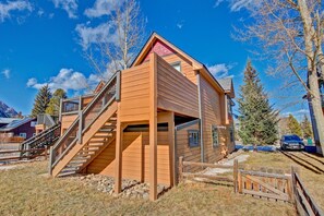 Eine zweistöckige Holzhütte mit einer Treppe, die zu der oberen Etage führt, umgeben von Bäumen und wolkenlosem Himmel.