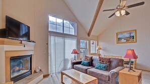 A cozy living room with a fireplace, a flat-screen TV, a ceiling fan, and a large window with curtains. - Tarn Landing  #c-4 2 Bedroom Condo by RedAwning (Frisco)