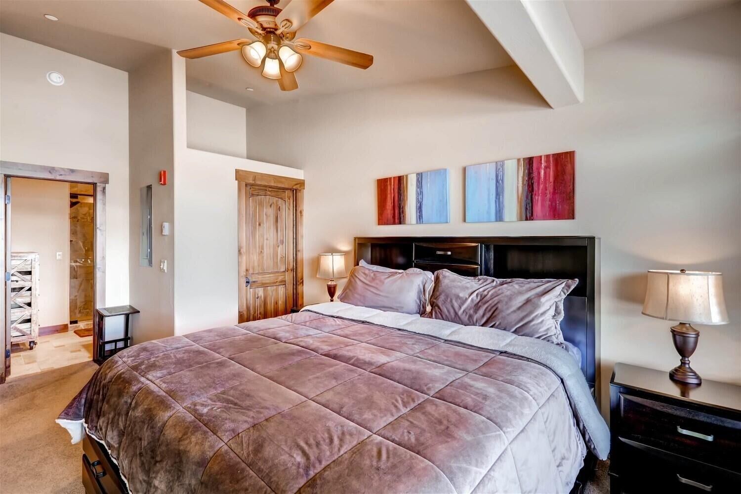 A bedroom with a large bed, a ceiling fan, a wooden door, and abstract wall art.