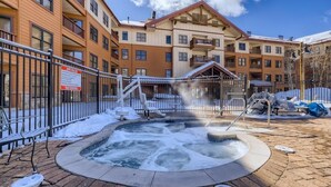 A spa area with a hot tub surrounded by a metal fence, a sign indicating spa rules, and a building with multiple balconies in the background. - Copper Springs  209 2 Bedroom Condo by RedAwning (Copper Mountain)