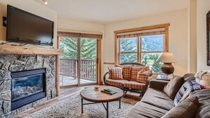A cozy living room with a stone fireplace, a flat-screen TV, a plaid sofa, a round coffee table, and a large window with a view of trees. - Copper Springs  209 2 Bedroom Condo by RedAwning (Copper Mountain)