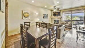 A spacious living room with a wooden dining table and chairs, a fireplace, and a large window offering a view of the outdoors. - S Off Main  #4 2 Bedroom Condo by RedAwning (Frisco)