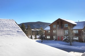 Un chalet en bois, dont le toit est recouvert de neige, doté de nombreuses fenêtres et d’une porte rouge, entouré de neige et d’arbres, avec des montagnes en arrière-plan.