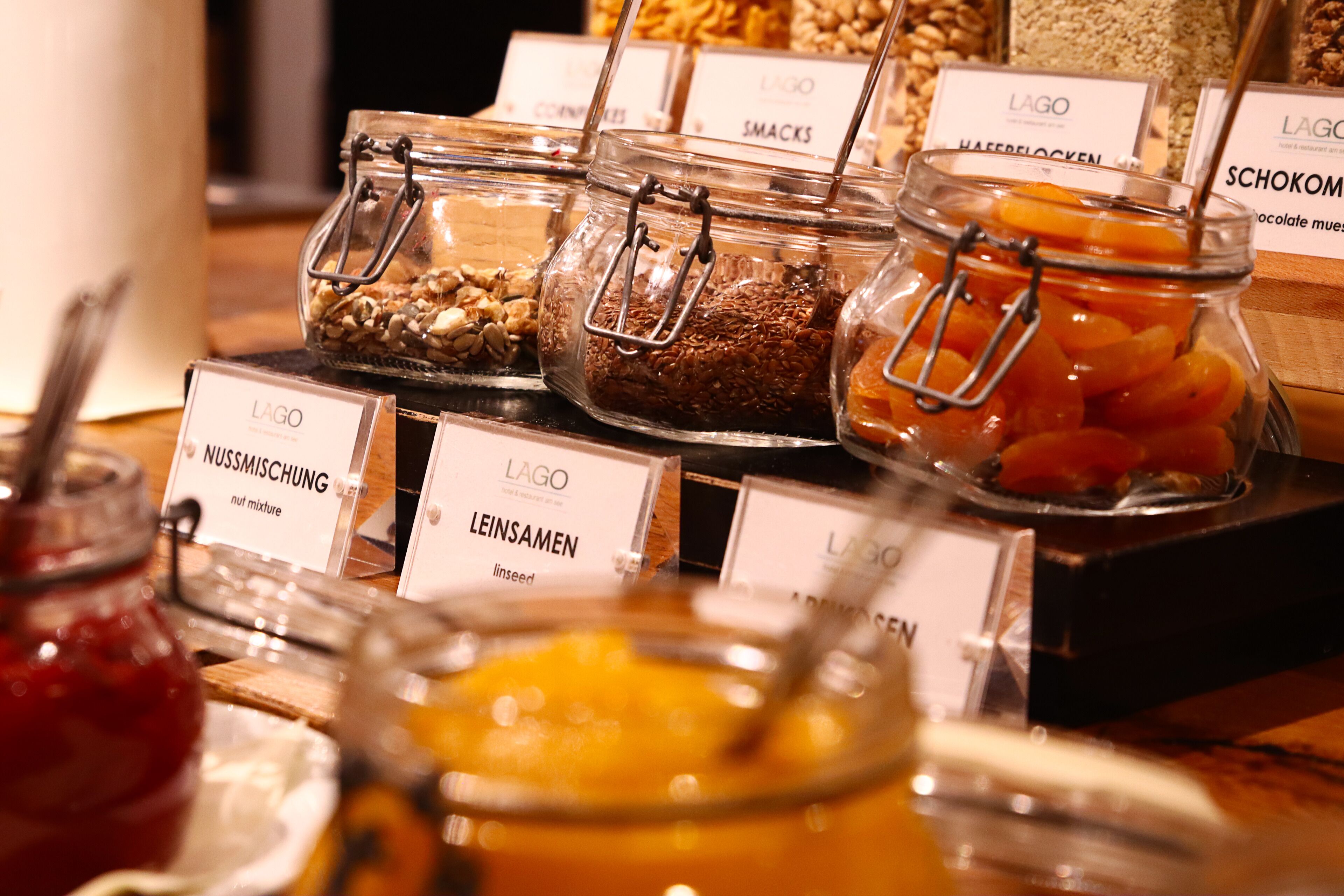 Variedad de alimentos en frascos de vidrio con etiquetas, como nueces, semillas y frutas deshidratadas.