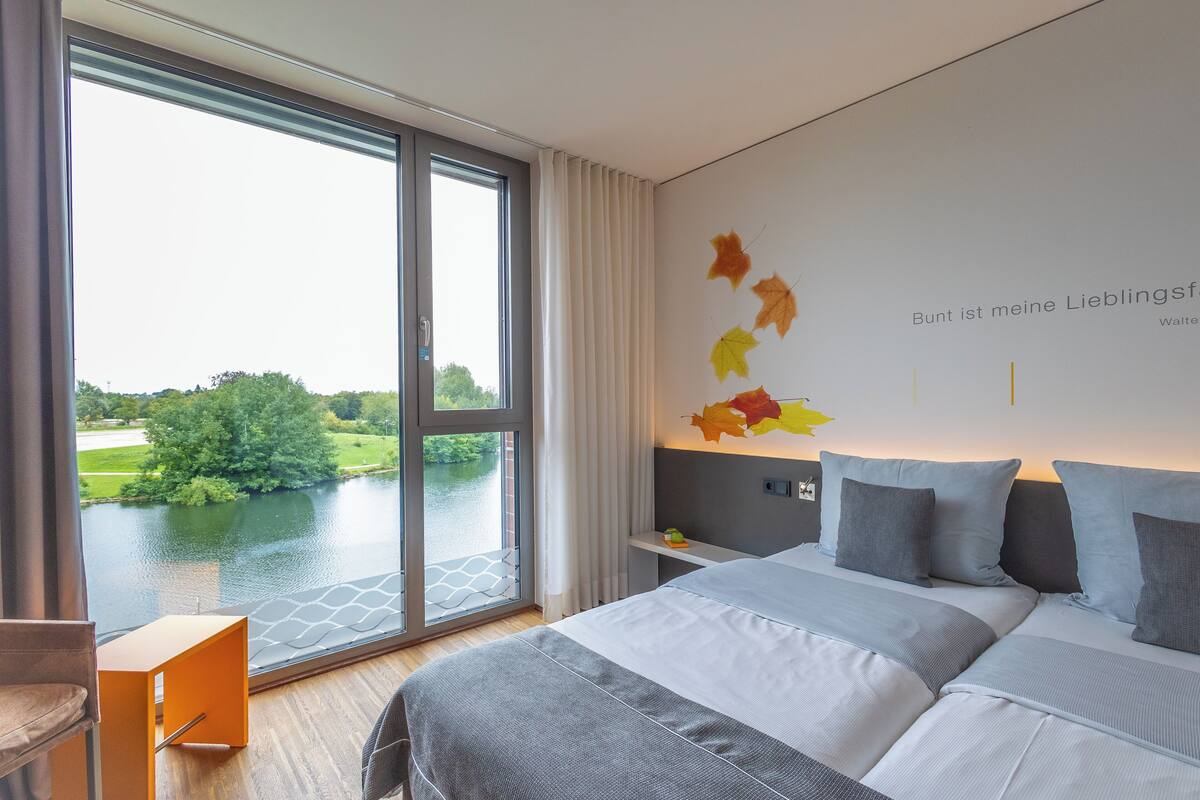 A modern hotel room with a large window, a bed with grey bedding, and autumn leaf decorations on the wall.