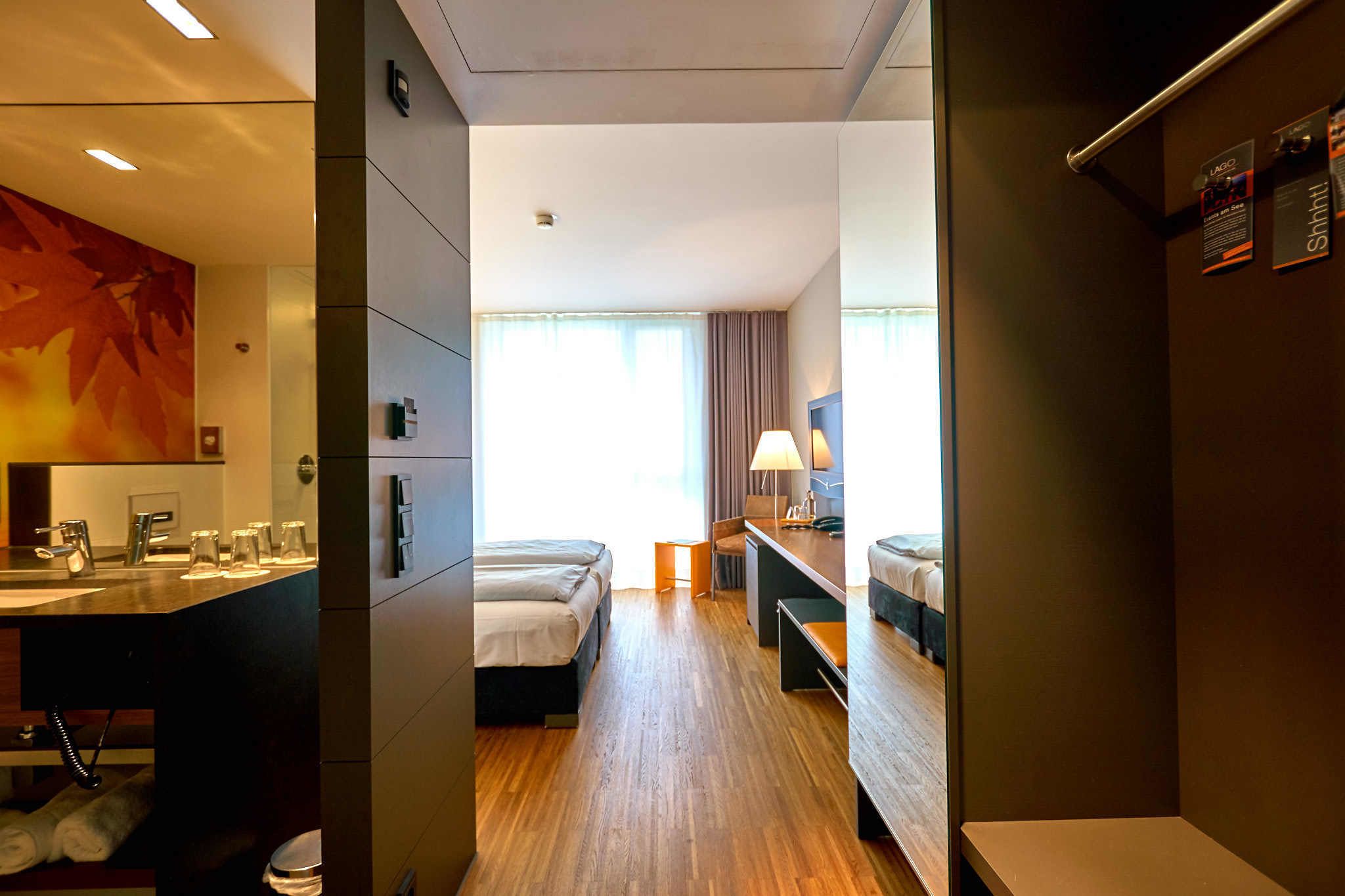 Habitación de hotel con piso de madera, un escritorio con lámpara, dos camas, baño con lavamanos y espejo, y un ventanal con cortinas.