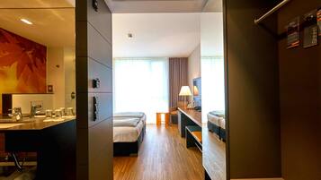 Habitación de hotel con piso de madera, un escritorio con lámpara, dos camas, baño con lavamanos y espejo, y un ventanal con cortinas.