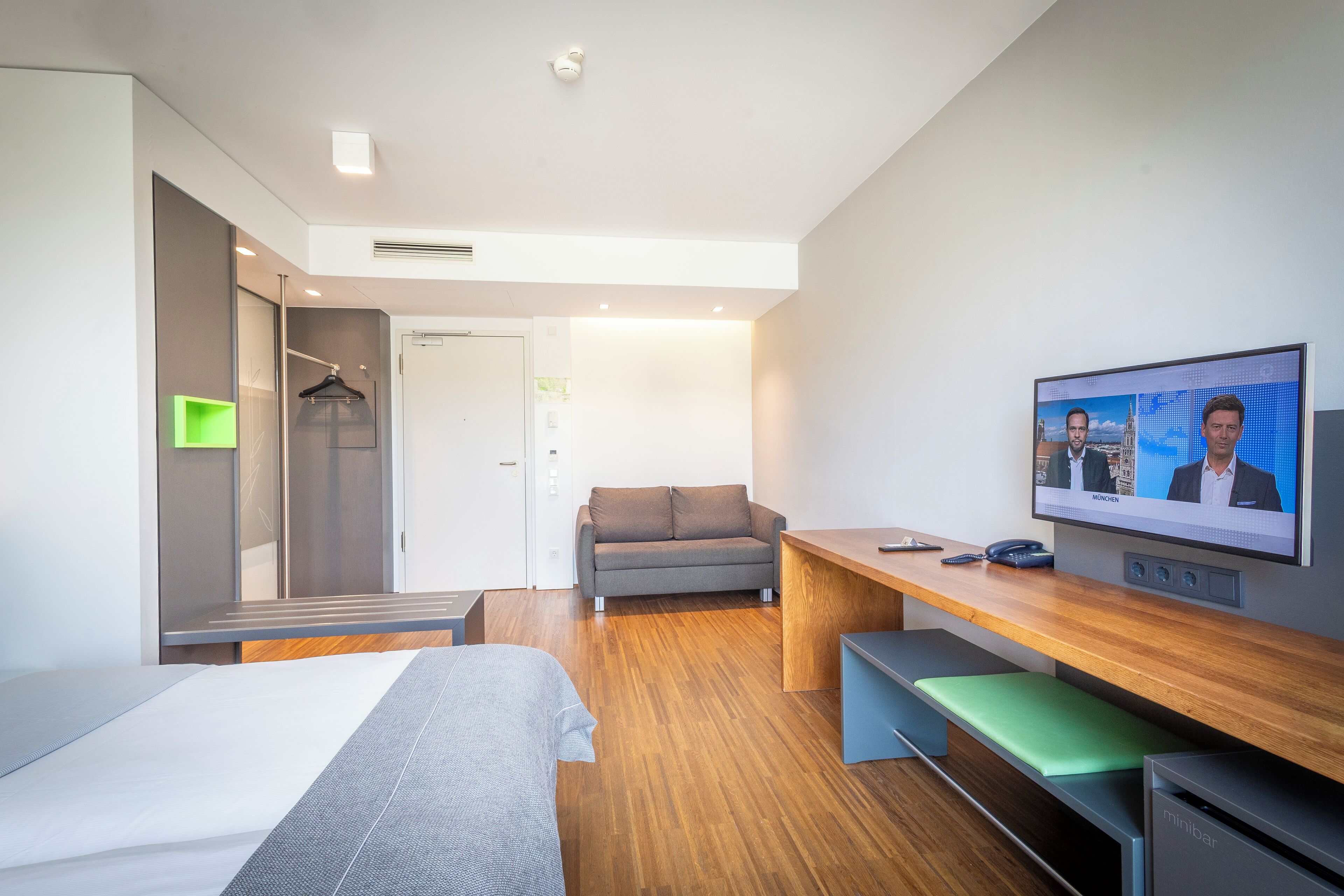 Ein modernes Hotelzimmer mit einem Bett, einem Sofa, einem Schreibtisch und einem Fernseher, der einen Videoanruf zeigt.