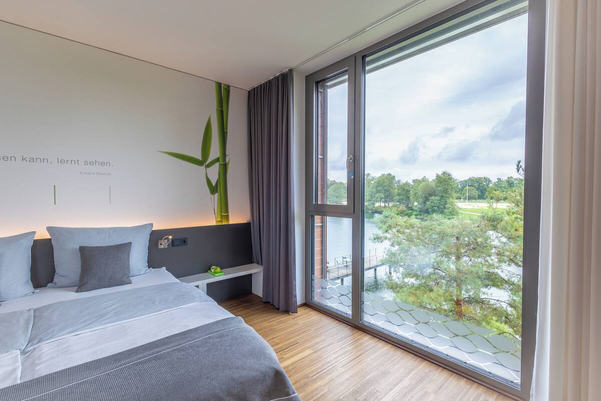 A modern bedroom with a large window, a bed with grey bedding, a bedside table, and a wall-mounted plant.
