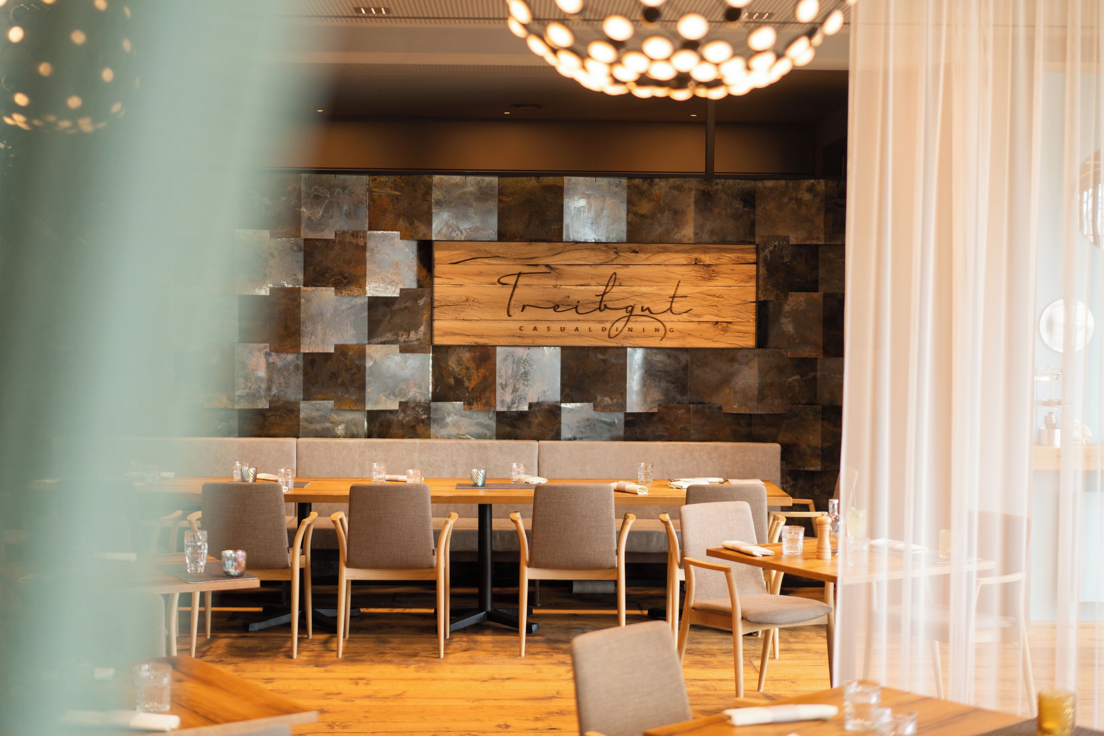 A restaurant with a wooden sign reading "Treibgut Casual Dining," featuring a modern interior with wooden tables and chairs, a stone wall, and a chandelier.