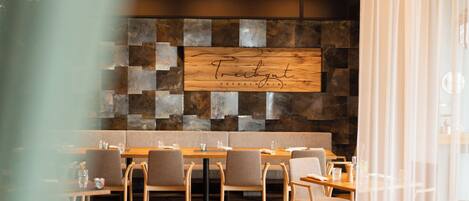 A restaurant with a wooden sign reading "Treibgut Casual Dining," featuring a modern interior with wooden tables and chairs, a stone wall, and a chandelier.
