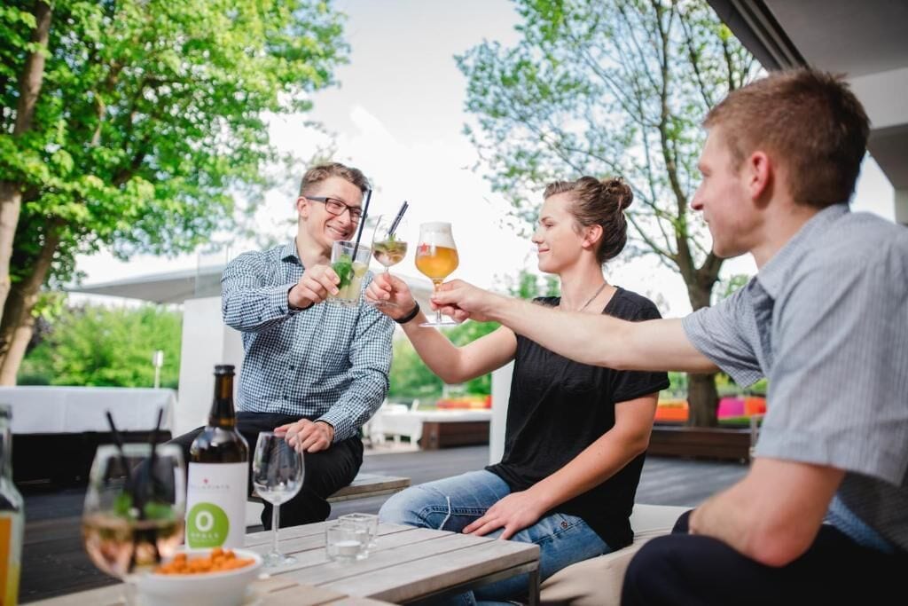 Un groupe de personnes trinquant avec des boissons dans un espace de restauration en plein air, avec en arrière-plan des arbres et un bâtiment.