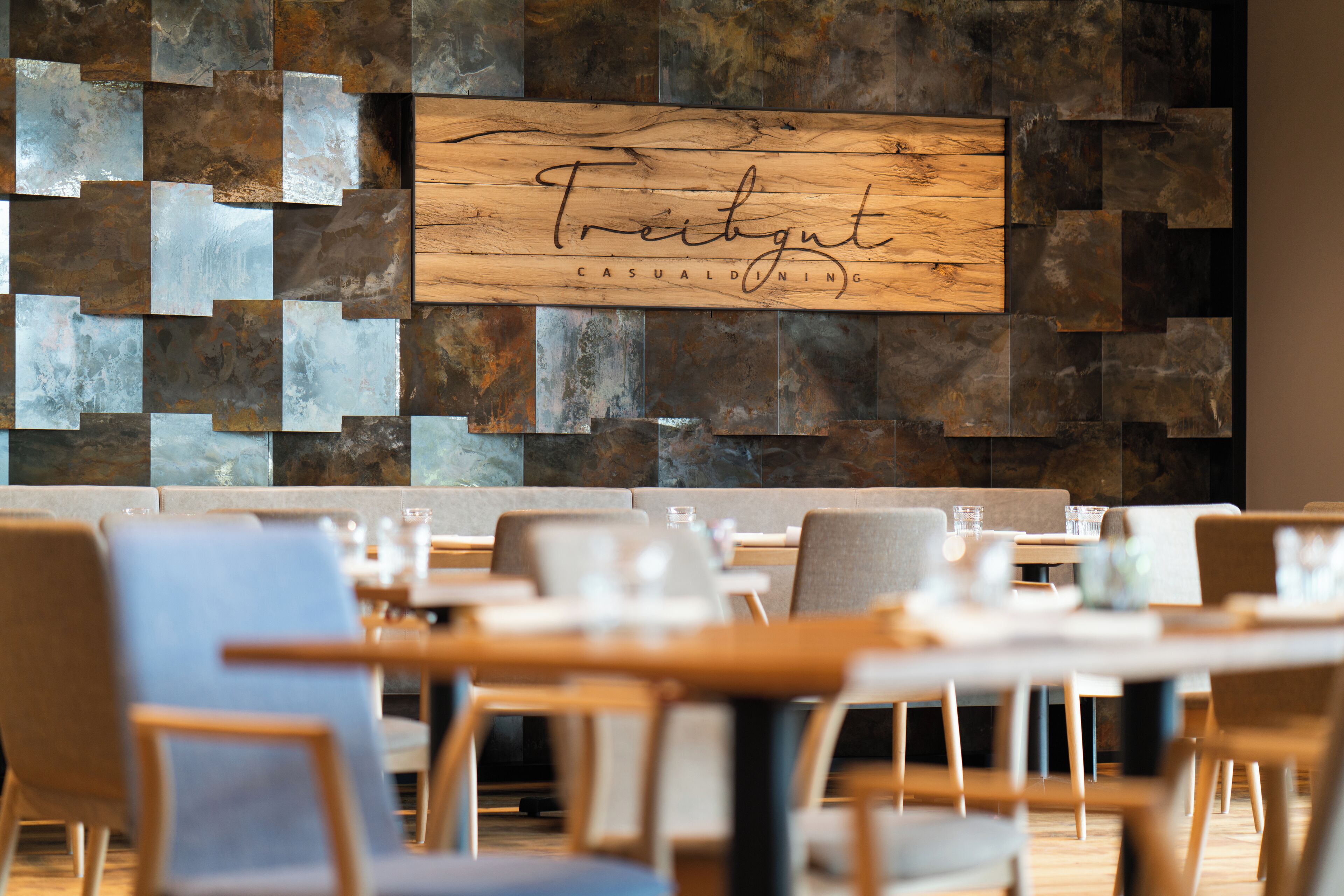 A restaurant interior with a wooden sign reading "Treibgut Casual Dining" and a modern, geometric wall design. Tables and chairs are arranged for dining.