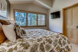 A bedroom with a large bed, a window with a forest view, a TV mounted on the wall, and a wooden door.